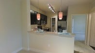 This single-story space showcases a thoughtfully designed open-concept kitchen that serves as a central feature of the layout. The kitchen is outfitted with rich, dark espresso cabinetry that extends to the upper walls, providing generous storage throughout. Stainless steel appliances, including a French door refrigerator, microwave, and range, lend a polished, contemporary feel to the space.

A wide peninsula breakfast bar with a light-toned stone countertop extends into the living area, creating a natural divide between the kitchen and adjoining spaces while offering casual seating and additional workspace. The bar is illuminated by two cylindrical pendant lights that hang at varying heights, complementing the track lighting mounted on the ceiling above the cooking area.

The neutral, warm-beige walls and light-toned flooring provide a clean backdrop that balances the darker cabinetry. A walk-in pantry or storage closet is visible in the background, adding practical functionality. The overall ambiance is modern and inviting, with a cohesive design aesthetic that emphasizes both style and usability throughout the kitchen and open living area.