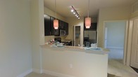 This single-story space showcases a thoughtfully designed open-concept kitchen that serves as a central feature of the layout. The kitchen is outfitted with rich, dark espresso cabinetry that extends to the upper walls, providing generous storage throughout. Stainless steel appliances, including a French door refrigerator, microwave, and range, lend a polished, contemporary feel to the space.

A wide peninsula breakfast bar with a light-toned stone countertop extends into the living area, creating a natural divide between the kitchen and adjoining spaces while offering casual seating and additional workspace. The bar is illuminated by two cylindrical pendant lights that hang at varying heights, complementing the track lighting mounted on the ceiling above the cooking area.

The neutral, warm-beige walls and light-toned flooring provide a clean backdrop that balances the darker cabinetry. A walk-in pantry or storage closet is visible in the background, adding practical functionality. The overall ambiance is modern and inviting, with a cohesive design aesthetic that emphasizes both style and usability throughout the kitchen and open living area.