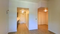 This single-floor space offers a clean, modern layout designed for comfortable everyday living. The interior is unified by warm light wood-toned flooring that flows seamlessly throughout, creating a cohesive and welcoming atmosphere. Flush-mount ceiling fixtures provide consistent, even lighting across all areas, complementing the bright white walls that lend the space an airy, open feel. A central hallway connects the main living areas to the entryway, which features a crisp white door and overhead lighting. To one side, the kitchen is visible with cabinetry, countertops, and appliances neatly arranged, offering a practical and efficient cooking space. The open-plan design allows the living area to flow naturally into adjoining spaces, with a clear sightline that enhances the sense of spaciousness. Neutral wall tones throughout provide a versatile backdrop suitable for a variety of interior styles. The layout is thoughtfully organized, with defined zones for living, cooking, and entry that remain visually connected. The overall ambiance is bright, clean, and functional, making this a well-appointed single-level space ideal for day-to-day living.