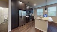 This single-floor residence offers a thoughtfully designed layout that blends modern aesthetics with functional living spaces. The open-concept kitchen and living area serves as the heart of the home, featuring rich dark wood cabinetry that extends to the ceiling, providing generous storage. A full suite of stainless steel appliances — including a side-by-side refrigerator with water dispenser, electric range, and over-the-range microwave — anchors the kitchen, which is further enhanced by a sleek horizontal tile backsplash and light stone countertops. A central island with a built-in sink and pendant lighting above adds both workspace and visual interest, while multiple windows fitted with horizontal blinds fill the area with natural light. The adjacent living space continues the rich hardwood-style flooring and is finished with a ceiling fan and access to an outdoor area. Bedrooms offer plush carpeting, natural light through blinds-fitted windows, and ample space. The bathrooms are well-finished, featuring dark floor-to-ceiling tile surrounds, stone-topped vanities with undermount sinks, and dark-framed mirrors. The primary bathroom includes a double vanity and a separate tiled shower. Neutral wall tones and recessed lighting throughout create a cohesive, contemporary ambiance.