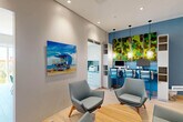 Located at 601 E Dania Beach Blvd in Dania Beach, Florida, this thoughtfully designed single-floor amenity space blends coastal aesthetics with modern functionality. The interior is unified by light wood-tone flooring, recessed lighting, and a crisp neutral color palette accented by bold ocean-inspired blues throughout.

The lounge area serves as a welcoming gathering space, furnished with mid-century modern teal upholstered chairs arranged around a natural wood coffee table. A vibrant Florida beach-themed canvas artwork adorns the wall, reinforcing the coastal atmosphere. Glass entry doors and open doorways connect the space seamlessly, allowing natural light to flow through.

A built-in business center station lines one wall, featuring individual computer workstations, blue bar-height chairs, and a striking living moss wall installation as a dramatic backdrop — a standout biophilic design element that adds texture and color.

The conference and dining area offers a sleek rectangular table surrounded by teal upholstered chairs beneath a sculptural wave-form pendant light fixture. A large wall-mounted display screen faces the table, making it ideal for presentations. Adjacent built-in white cabinetry with open shelving and deep blue accent surfaces provides ample storage, while a connected kitchenette with a vibrant teal backsplash offers convenient service support. Together, these areas create a cohesive, professional, and inviting environment.