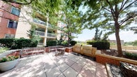 This single-floor space offers a functional and well-appointed layout that connects clean, carpeted interior corridors to a standout outdoor amenity area. The interior hallways feature neutral gray walls, patterned carpet tile flooring in earthy tones, and glass-paneled doors with frosted signage, including access to a designated "Zen Garden" area. The corridors are practical and straightforward, with safety fixtures, a built-in display panel, and glass sidelights that draw in views of the surrounding outdoor environment.

The highlight of the space is the expansive outdoor patio, which is thoughtfully designed for both relaxation and entertaining. Large-format tile pavers provide a clean, durable surface underfoot, while mature trees offer generous natural shade across the area. A substantial built-in outdoor kitchen anchors one side of the patio, featuring multiple professional-grade grills and a stone-and-brick surround that speaks to quality construction. Wooden benches and casual seating arrangements are integrated throughout, creating inviting social spaces. Neatly trimmed hedges and lush greenery frame the perimeter, enhancing the sense of privacy and tranquility. The patio sits adjacent to a multi-story residential building, completing the communal outdoor experience.