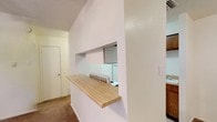 This single-story space showcases a practical and efficiently designed interior with a notable kitchen pass-through as its defining architectural feature. The pass-through opening connects the kitchen to the adjacent living area, framed by clean white walls and topped with a warm-toned butcher block countertop that serves as a breakfast bar. This design choice promotes an open, communicative feel between the two spaces while maintaining a degree of separation.

The kitchen, visible through the pass-through, features white appliances, including a refrigerator, and is complemented by warm wood-toned cabinetry on the right side. The combination of white walls and wood elements creates a balanced, neutral aesthetic throughout.

The flooring in the living area consists of carpeting in a neutral tone, contributing to a comfortable, casual atmosphere. White walls throughout keep the space feeling bright and clean. A textured ceiling adds a subtle layer of visual detail. A hallway leading to additional rooms is visible in the background, hinting at the broader layout of the unit. Overall, the space presents a functional and straightforward design suited for everyday living.