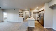 This single-floor residence offers a thoughtfully designed layout that balances open living with functional private spaces. The heart of the home is the open-concept kitchen and living area, where wood-look flooring transitions seamlessly into plush carpeting. The kitchen is equipped with stainless steel appliances, gray shaker-style cabinetry, light speckled stone countertops, and a central island with a built-in sink — ideal for both cooking and casual dining. A built-in desk nook and open floating shelves along the adjacent wall add practical character to the space. The living area is generously sized, featuring a ceiling fan and natural light from large windows with blinds. A hallway connects the living spaces to the private bedrooms, each finished with soft carpeting, ceiling fans, and windows that allow ample natural light. The bedrooms include closet space, with at least one room featuring a walk-in closet with built-in shelving. The bathrooms are finished with wood-look flooring, gray cabinetry, stone-topped vanities, and tiled tub-shower combinations. A neutral color palette throughout lends the space a clean, modern aesthetic.