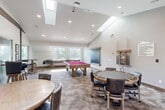 Located at 1600 N 1575 W in Layton, Utah, this single-floor entertainment space is designed to deliver an exceptional social and recreational experience. The layout is divided into two distinct living areas and a kitchenette, each thoughtfully appointed for comfort and function.

The centerpiece of the space is a dedicated home theater room featuring vaulted ceilings, a ceiling-mounted projector, a large projection screen, and a soundbar. Rows of plush, dark leather reclining seats with built-in cup holders and armrest consoles line the carpeted room, which is accented by framed classic movie posters. Natural light filters in through windows along the side wall, complementing the recessed lighting overhead.

The second living area serves as a versatile game and lounge room, highlighted by soaring vaulted ceilings with exposed dark wood beam accents. This open area accommodates a pool table, a shuffleboard table, and multiple seating arrangements including a round dining table and bar-height stools, creating an inviting gathering space. Large windows throughout flood the room with natural light and offer views of the surrounding greenery.

The kitchenette is finished with rich, dark wood cabinetry, stone-look tile flooring, stainless steel appliances, and a window above the sink. A distinctive rustic barn-style frosted glass door adds a stylish architectural touch, seamlessly connecting the kitchen to the rest of the space.