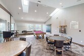Located at 1600 N 1575 W in Layton, Utah, this single-floor entertainment space is designed to deliver an exceptional social and recreational experience. The layout is divided into two distinct living areas and a kitchenette, each thoughtfully appointed for comfort and function.

The centerpiece of the space is a dedicated home theater room featuring vaulted ceilings, a ceiling-mounted projector, a large projection screen, and a soundbar. Rows of plush, dark leather reclining seats with built-in cup holders and armrest consoles line the carpeted room, which is accented by framed classic movie posters. Natural light filters in through windows along the side wall, complementing the recessed lighting overhead.

The second living area serves as a versatile game and lounge room, highlighted by soaring vaulted ceilings with exposed dark wood beam accents. This open area accommodates a pool table, a shuffleboard table, and multiple seating arrangements including a round dining table and bar-height stools, creating an inviting gathering space. Large windows throughout flood the room with natural light and offer views of the surrounding greenery.

The kitchenette is finished with rich, dark wood cabinetry, stone-look tile flooring, stainless steel appliances, and a window above the sink. A distinctive rustic barn-style frosted glass door adds a stylish architectural touch, seamlessly connecting the kitchen to the rest of the space.