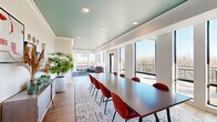 Located at 730 1/2 N First St in Minneapolis's vibrant North Loop neighborhood, this single-floor amenity space offers a thoughtfully designed interior that blends functionality with modern style. The layout encompasses a combined dining and living room along with a connecting hallway, making efficient use of its footprint.

The dining area is anchored by a large dark conference-style table surrounded by terracotta-hued upholstered chairs, set atop a textured area rug. A wall-mounted flat-screen TV is framed by decorative botanical wallcovering, while warm wood cabinetry and curated wall art add character and depth. Lush indoor plants contribute a fresh, organic element throughout.

The living area transitions seamlessly from the dining space and features floor-to-ceiling windows on multiple walls, flooding the room with natural light and offering sweeping views of the Minneapolis skyline and surrounding treetops. A glass-paneled door provides direct access to an outdoor terrace, further blurring the boundary between interior and exterior. Comfortable lounge seating, a geometric navy area rug, and a modern chandelier complete the inviting atmosphere.

Throughout the space, a signature teal-painted ceiling creates a cohesive and eye-catching design element, complemented by crisp white walls and light wood-tone flooring. Recessed lighting ensures consistent illumination across both areas.