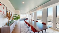 Located at 730 1/2 N First St in Minneapolis's vibrant North Loop neighborhood, this single-floor amenity space offers a thoughtfully designed interior that blends functionality with modern style. The layout encompasses a combined dining and living room along with a connecting hallway, making efficient use of its footprint.

The dining area is anchored by a large dark conference-style table surrounded by terracotta-hued upholstered chairs, set atop a textured area rug. A wall-mounted flat-screen TV is framed by decorative botanical wallcovering, while warm wood cabinetry and curated wall art add character and depth. Lush indoor plants contribute a fresh, organic element throughout.

The living area transitions seamlessly from the dining space and features floor-to-ceiling windows on multiple walls, flooding the room with natural light and offering sweeping views of the Minneapolis skyline and surrounding treetops. A glass-paneled door provides direct access to an outdoor terrace, further blurring the boundary between interior and exterior. Comfortable lounge seating, a geometric navy area rug, and a modern chandelier complete the inviting atmosphere.

Throughout the space, a signature teal-painted ceiling creates a cohesive and eye-catching design element, complemented by crisp white walls and light wood-tone flooring. Recessed lighting ensures consistent illumination across both areas.