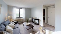 This single-floor residential unit offers a comfortable and thoughtfully designed layout across several distinct spaces. The living room is a welcoming area finished with plush carpeting and painted in a soft, cool gray tone. Natural light filters in through large windows dressed with sheer white curtains, creating a bright and airy atmosphere. A wall-unit HVAC system ensures year-round comfort, while decorative wall art adds a stylish touch to the space.

The kitchen features sleek gray cabinetry paired with light marble-patterned countertops and a classic subway tile backsplash. A freestanding electric coil range anchors the cooking area, providing a fully functional setup for everyday meal preparation.

Both bedrooms are carpeted and share a striking design feature: a bold, full-wall exposed brick accent that adds warmth, texture, and character to each room. The bedrooms are generously proportioned and include windows that bring in natural light. One bedroom also features a sliding-door closet with built-in organization, offering practical storage solutions.

The bathroom is clean and bright, with wood-look flooring, a full shower-tub combination with white tile surround, and a crisp white vanity. The overall ambiance of this unit blends modern updates with distinctive architectural details, resulting in a cozy yet stylish living environment.