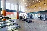 This expansive fitness center is designed to accommodate a comprehensive range of workout disciplines across a generously sized single-floor layout. The space is divided into distinct functional zones, each thoughtfully appointed with high-quality equipment and purposeful design elements.

The main cardio and strength area commands attention with soaring double-height ceilings clad in warm wood paneling, punctuated by industrial-style pendant lighting and large floor-to-ceiling windows that flood the space with natural light. True-brand treadmills, ellipticals, stair climbers, and cable machines are arranged throughout this open zone, which overlooks an outdoor pool area.

A dedicated functional training studio features light hardwood flooring, a teal-painted ceiling, floor-to-ceiling glass walls, TRX suspension systems, spin bikes, stability balls, and medicine balls. Large sliding glass doors connect this studio directly to an outdoor artificial turf area equipped with sleds, dumbbells, and additional functional training tools under open sky.

A support corridor provides a hand-washing station with decorative tiled accents and a dumbbell rack. The overall aesthetic blends contemporary finishes—teal ceilings, geometric wall treatments, and warm column details—creating a cohesive, energetic environment suited for diverse fitness activities.