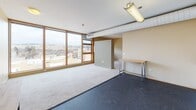 Located in Ithaca, NY, this single-floor unit offers a thoughtfully laid-out interior with a mix of practical finishes and striking views. The main living area is generously proportioned, featuring a combination of dark resilient flooring and plush carpeting that defines distinct zones within the open layout. Large wood-framed windows span a full wall, flooding the space with natural light and offering sweeping elevated views of the Ithaca cityscape and surrounding hillside landscape. Exposed ductwork along the ceiling adds an industrial character to the space, complementing the warm beige walls throughout.

The kitchen is efficiently designed with white upper and lower cabinetry, dark countertops, and a full suite of white appliances including an electric range, dishwasher, and refrigerator. An overhead ventilation hood and a stainless steel sink complete the functional setup.

The private bathroom features dark slate-style tile flooring, a walk-in shower, a wall-mounted vanity with an integrated white basin, a large mirror with overhead lighting, and clean, minimal finishes throughout.

Additionally, a shared restroom facility is accessible within the building, equipped with multiple stalls, fixtures, and large windows providing natural light. The overall aesthetic of the space is clean, functional, and urban.