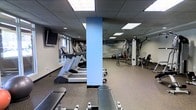 This fitness center is a single-level space thoughtfully designed to accommodate a comprehensive workout experience. The room features a dropped ceiling with recessed panel lighting that evenly illuminates the entire area, creating a bright and energizing atmosphere. Durable rubber flooring in a speckled finish covers the space, providing both safety and comfort underfoot.

The cardio zone is well-appointed with multiple treadmills and an elliptical machine, all positioned near a window that allows natural light to filter in. Wall-mounted televisions are installed nearby to enhance the workout experience. A sturdy structural column, finished in a light blue accent, serves as a natural divider between the cardio and strength training areas.

The strength training section houses a multi-station cable and weight machine, a rowing machine, and a variety of free weights including dumbbells and a medicine ball, offering users a full range of resistance training options. Workout benches are also available throughout the space.

A standout feature is the motivational text wall graphic, displaying words such as "strength," "dedication," "focus," and "improve," reinforcing an encouraging and goal-oriented atmosphere. The overall design is clean, functional, and purpose-built to support an active lifestyle.