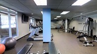 This fitness center is a single-level space thoughtfully designed to accommodate a comprehensive workout experience. The room features a dropped ceiling with recessed panel lighting that evenly illuminates the entire area, creating a bright and energizing atmosphere. Durable rubber flooring in a speckled finish covers the space, providing both safety and comfort underfoot.

The cardio zone is well-appointed with multiple treadmills and an elliptical machine, all positioned near a window that allows natural light to filter in. Wall-mounted televisions are installed nearby to enhance the workout experience. A sturdy structural column, finished in a light blue accent, serves as a natural divider between the cardio and strength training areas.

The strength training section houses a multi-station cable and weight machine, a rowing machine, and a variety of free weights including dumbbells and a medicine ball, offering users a full range of resistance training options. Workout benches are also available throughout the space.

A standout feature is the motivational text wall graphic, displaying words such as "strength," "dedication," "focus," and "improve," reinforcing an encouraging and goal-oriented atmosphere. The overall design is clean, functional, and purpose-built to support an active lifestyle.