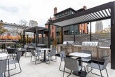 Located at 137 N West Temple in the heart of Salt Lake City, this thoughtfully designed outdoor terrace offers a refined and functional amenity space set against a striking contemporary brick building. The terrace is finished with large-format light stone pavers that lend a clean, upscale aesthetic throughout. Two sleek dark-framed pergolas anchor the space, one of which shelters a fully equipped outdoor kitchen featuring stainless steel grills and a stone-clad counter — ideal for entertaining and communal gatherings. Scattered across the terrace are multiple seating arrangements, including woven lounge chairs, café-style tables, and elevated bar-height seating, accommodating a variety of social settings. A central fire pit with river rock fill provides a cozy focal point for relaxed gatherings. Horizontal slatted privacy screens and ornamental fencing define the perimeter while maintaining an open feel. The terrace adjoins the building directly through full-height glass sliding and double doors, seamlessly connecting indoor and outdoor spaces. Views of the surrounding Salt Lake City skyline and neighboring historic architecture add to the urban character of the space, making this terrace a compelling outdoor destination in a vibrant downtown setting.