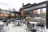 Located at 137 N West Temple in the heart of Salt Lake City, this thoughtfully designed outdoor terrace offers a refined and functional amenity space set against a striking contemporary brick building. The terrace is finished with large-format light stone pavers that lend a clean, upscale aesthetic throughout. Two sleek dark-framed pergolas anchor the space, one of which shelters a fully equipped outdoor kitchen featuring stainless steel grills and a stone-clad counter — ideal for entertaining and communal gatherings. Scattered across the terrace are multiple seating arrangements, including woven lounge chairs, café-style tables, and elevated bar-height seating, accommodating a variety of social settings. A central fire pit with river rock fill provides a cozy focal point for relaxed gatherings. Horizontal slatted privacy screens and ornamental fencing define the perimeter while maintaining an open feel. The terrace adjoins the building directly through full-height glass sliding and double doors, seamlessly connecting indoor and outdoor spaces. Views of the surrounding Salt Lake City skyline and neighboring historic architecture add to the urban character of the space, making this terrace a compelling outdoor destination in a vibrant downtown setting.