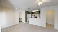 This single-floor space features an open-concept design that seamlessly connects the living area to the kitchen. The living room is finished with neutral carpet, white baseboards, and crown molding details, creating a clean and cohesive aesthetic. A ceiling fan with an integrated light fixture provides both comfort and illumination, while a track lighting system along the adjacent wall offers additional, adjustable lighting options.

The kitchen is positioned behind a raised breakfast bar counter, providing a natural division between the cooking and living spaces while maintaining an open feel. Light wood cabinetry lines the upper walls of the kitchen, complemented by white appliances including a refrigerator and microwave. A fluorescent overhead fixture ensures the kitchen is well-lit. The darker accent wall within the kitchen adds a subtle contrast to the otherwise neutral color palette throughout the space.

A distinctive arched entryway leads from the living area into a hallway or additional room, adding an architectural touch that enhances the overall character of the unit. The consistent use of white walls and light-toned flooring materials throughout contributes to a bright, airy atmosphere.