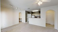 This single-floor space features an open-concept design that seamlessly connects the living area to the kitchen. The living room is finished with neutral carpet, white baseboards, and crown molding details, creating a clean and cohesive aesthetic. A ceiling fan with an integrated light fixture provides both comfort and illumination, while a track lighting system along the adjacent wall offers additional, adjustable lighting options.

The kitchen is positioned behind a raised breakfast bar counter, providing a natural division between the cooking and living spaces while maintaining an open feel. Light wood cabinetry lines the upper walls of the kitchen, complemented by white appliances including a refrigerator and microwave. A fluorescent overhead fixture ensures the kitchen is well-lit. The darker accent wall within the kitchen adds a subtle contrast to the otherwise neutral color palette throughout the space.

A distinctive arched entryway leads from the living area into a hallway or additional room, adding an architectural touch that enhances the overall character of the unit. The consistent use of white walls and light-toned flooring materials throughout contributes to a bright, airy atmosphere.