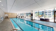 Located at 218 W Lake St in Chicago's vibrant Loop neighborhood, this indoor aquatic facility offers a refined and well-appointed environment designed for both fitness and relaxation. The centerpiece is a multi-lane lap pool with crystal-clear blue water, illuminated by recessed underwater lighting and overhead can lights that create a bright, inviting atmosphere throughout the space.

The pool area is framed by richly textured slate-tiled walls in cool gray tones, complemented by warm, stacked-stone accent walls that add visual depth and warmth to the interior. The smooth, paneled ceiling enhances the sense of openness within the room. Safety amenities, including handrails, a life ring, and first aid provisions, are thoughtfully integrated into the design.

A standout feature is the dramatic wall of floor-to-ceiling glass along one side of the pool, which opens visually and physically to an outdoor terrace. The terrace is landscaped with vibrant planters, lounge seating, outdoor dining furniture, and a fire feature, all set against a striking backdrop of Chicago's downtown high-rise skyline. This seamless indoor-outdoor connection fills the space with natural light and urban panoramic views. Stone-tiled flooring and restroom access complete the functional, polished amenity space.
