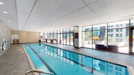 Located at 218 W Lake St in Chicago's vibrant Loop neighborhood, this indoor aquatic facility offers a refined and well-appointed environment designed for both fitness and relaxation. The centerpiece is a multi-lane lap pool with crystal-clear blue water, illuminated by recessed underwater lighting and overhead can lights that create a bright, inviting atmosphere throughout the space.

The pool area is framed by richly textured slate-tiled walls in cool gray tones, complemented by warm, stacked-stone accent walls that add visual depth and warmth to the interior. The smooth, paneled ceiling enhances the sense of openness within the room. Safety amenities, including handrails, a life ring, and first aid provisions, are thoughtfully integrated into the design.

A standout feature is the dramatic wall of floor-to-ceiling glass along one side of the pool, which opens visually and physically to an outdoor terrace. The terrace is landscaped with vibrant planters, lounge seating, outdoor dining furniture, and a fire feature, all set against a striking backdrop of Chicago's downtown high-rise skyline. This seamless indoor-outdoor connection fills the space with natural light and urban panoramic views. Stone-tiled flooring and restroom access complete the functional, polished amenity space.