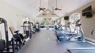 This fitness center at 850 S Cimarron Way in Aurora, Colorado, offers a well-equipped, single-floor exercise facility designed to support a full range of workout routines. The space is defined by its striking vaulted ceilings, which lend an open, airy feel and are complemented by multiple ceiling fans that ensure consistent air circulation throughout. A row of arched windows along one side floods the interior with natural light, while slatted blinds on the opposing windows provide adjustable brightness control.

The facility is outfitted with a comprehensive selection of cardio machines, including treadmills, ellipticals, and a stair-stepper, all positioned along one side of the floor. On the opposite side, a variety of strength and resistance training machines are arranged for easy access, alongside a full dumbbell rack offering a complete range of free weights. A large wall-to-wall mirror along one end of the room visually expands the space and allows users to monitor their form during exercise.

The flooring throughout is durable rubber gym matting, providing both safety and comfort underfoot. A wall-mounted flat-screen television adds convenience for users during cardio sessions. French doors at the rear of the space lead to an adjacent area, adding connectivity and accessibility. Recessed lighting and neutral wall tones complete the clean, functional aesthetic of this well-maintained fitness facility.