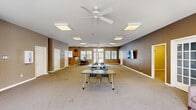 Located at 2415 SE Fourth Lane in Gainesville, Florida, this single-floor community space offers a functional and welcoming layout designed for group use. The main living area is expansive and open, featuring warm taupe-toned walls, neutral carpet flooring, and a wall-mounted flat-screen television. A set of elegant French doors leads toward additional rooms, while large front-facing windows and glass entry doors flood the space with natural light and provide views of the surrounding outdoor area. Ceiling fans and flush-mounted lighting fixtures contribute to a comfortable and well-lit atmosphere throughout.

The kitchen is equipped with warm wood cabinetry, granite-style countertops, a double stainless steel sink, and a white range with overhead ventilation. A peninsula counter separates the kitchen from an adjacent dining area, which features tile flooring and a large dining table with seating. Natural light enters through multiple windows fitted with blinds, enhancing the functionality of this shared cooking and dining space.

A secondary room serves as a dedicated reading or resource area, lined with tall bookshelves stocked with a wide variety of books. Carpeted and quietly lit with overhead fixtures, this room provides a calm, focused environment for individual study or group learning. Together, these areas create a versatile and community-oriented space well-suited for gatherings, programming, and everyday use.