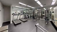 This fitness center is a comprehensively outfitted exercise facility designed to support a full range of workout routines. The space is laid out across a single floor with a practical, open floor plan that maximizes usable exercise area. Durable rubber flooring in a dark speckled finish covers the entire space, providing both safety and comfort underfoot during workouts.

The cardio zone features multiple Matrix-brand treadmills and elliptical trainers arranged in a row, all equipped with digital display consoles. A large wall of floor-to-ceiling mirrors lines one side of the room, visually expanding the space and allowing users to monitor their form during exercise.

The strength training area is well-appointed with a multi-station cable machine system, a leg press machine, and a flat weight bench, offering a variety of resistance training options. Framed wall art adds a touch of character to the otherwise clean, neutral aesthetic of the room.

Overhead, flush-mounted fluorescent panel lighting provides bright, even illumination throughout. A seating bench is positioned along one wall, offering a convenient rest area. The space also includes a wall-mounted device, likely a television, and a waste receptacle for user convenience. The overall ambiance is clean, functional, and conducive to focused fitness training.