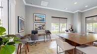 Located at 200 S Mallory St in Hampton, Virginia, this single-floor office space offers a thoughtfully designed environment that balances collaborative and individual work needs. The open layout is anchored by a large central conference-style table with upholstered bar-height seating, making it ideal for group meetings or shared work sessions. A wall-mounted flat-screen television and wall sconces lend a polished, professional atmosphere, while crown molding and soft blue-gray walls add architectural character and a calming aesthetic.

Rich wood cabinetry with granite countertops lines one wall, providing functional storage and surface space alongside a printer station. Light wood-look tile flooring runs throughout, complemented by a vibrant, multi-toned area rug that adds warmth and visual interest. Individual workstations equipped with desktop computers and bar-height seating are positioned near large windows fitted with roller shades, offering views of the surrounding greenery while allowing for natural light control.

Comfortable lounge seating is tucked into windowed nooks, creating inviting spots for focused work or casual conversation. Local artwork adorns the walls, giving the space a distinct sense of place. French doors open to additional areas, enhancing the sense of openness and connectivity throughout this well-curated, modern office environment.