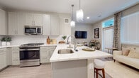 This well-appointed single-floor residence showcases a thoughtfully designed open-concept layout with a bright, neutral color palette and light wood-tone flooring that flows seamlessly throughout. At the heart of the home is a stylish kitchen featuring soft gray shaker-style cabinetry, a white subway tile backsplash, and a generous peninsula island topped with striking veined quartz countertops. The island doubles as a casual dining space with bar seating, while geometric pendant lights above add a contemporary touch. Stainless steel appliances — including a gas range, built-in microwave, refrigerator, and dishwasher — complete this functional and elegant kitchen. The space opens naturally into the living and dining areas, creating a connected, sociable environment with access to natural light from nearby windows. The primary bedroom offers a serene retreat with a large window, warm neutral tones, and access to a spacious walk-in closet outfitted with multiple hanging rods and shelving on three walls. A second bedroom provides additional flexibility, also featuring a window with natural light. The bathroom is finished with a white vanity, quartz countertop, large mirror, and wood-look flooring, maintaining the home's clean and cohesive aesthetic throughout.