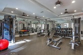 Located at 100 Drexel Point in Dunwoody, Georgia, this fitness center is a comprehensively outfitted facility designed to support a full range of workout routines. The space is organized across several interconnected zones, each dedicated to a specific type of training, and is unified by elegant architectural details including tray ceilings with crown molding and recessed lighting throughout.

The strength training area is the centerpiece of the facility, featuring a multi-station cable machine, a full rack of dumbbells, and multiple adjustable weight benches. Floor-to-ceiling mirrors line the walls, amplifying the sense of space and allowing users to monitor their form. Ceiling fans provide additional ventilation and comfort during intense sessions.

A separate cardio zone houses treadmills, stationary bikes, and other aerobic equipment, benefiting from generous natural light streaming through large windows. A dedicated spin bike area, equipped with a wall-mounted screen, offers an immersive cycling experience adjacent to a glass-paneled door that opens directly to an outdoor patio, blending indoor and outdoor environments.

The entry corridor connects these zones seamlessly and includes a hand sanitizer station and space for functional training accessories such as stability balls. A dramatic dark-wood staircase with wrought-iron balusters serves as a striking architectural focal point, while durable rubber flooring throughout ensures safety and functionality.