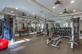Located at 100 Drexel Point in Dunwoody, Georgia, this fitness center is a comprehensively outfitted facility designed to support a full range of workout routines. The space is organized across several interconnected zones, each dedicated to a specific type of training, and is unified by elegant architectural details including tray ceilings with crown molding and recessed lighting throughout.

The strength training area is the centerpiece of the facility, featuring a multi-station cable machine, a full rack of dumbbells, and multiple adjustable weight benches. Floor-to-ceiling mirrors line the walls, amplifying the sense of space and allowing users to monitor their form. Ceiling fans provide additional ventilation and comfort during intense sessions.

A separate cardio zone houses treadmills, stationary bikes, and other aerobic equipment, benefiting from generous natural light streaming through large windows. A dedicated spin bike area, equipped with a wall-mounted screen, offers an immersive cycling experience adjacent to a glass-paneled door that opens directly to an outdoor patio, blending indoor and outdoor environments.

The entry corridor connects these zones seamlessly and includes a hand sanitizer station and space for functional training accessories such as stability balls. A dramatic dark-wood staircase with wrought-iron balusters serves as a striking architectural focal point, while durable rubber flooring throughout ensures safety and functionality.