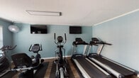This fitness center is a comprehensively outfitted single-floor exercise facility designed to support a wide range of workout routines. The space features calming blue-gray walls, durable wood-look flooring, and a textured white ceiling with crown molding accents, creating a clean and functional environment throughout.

The cardio zone is prominently featured and includes multiple treadmills by Spirit Fitness, an elliptical trainer, a stationary upright bike, and a spin bike, all positioned for easy use and accompanied by wall-mounted flat-screen televisions to keep users entertained during workouts. A ceiling-mounted linear light fixture ensures bright, even illumination across this area.

The strength training section houses a multi-station cable machine, an adjustable weight bench, and Inflight Fitness leg press and extension machines, providing ample options for resistance-based workouts. Protective rubber mats are placed beneath equipment throughout the space.

Functional training accessories are also available, including a rack of medicine balls and stability balls in various sizes. Natural light filters in through a window fitted with blinds, and a wall-mounted fan provides additional air circulation. The facility includes a dedicated entry door and posted fitness center rules, reflecting a well-managed and user-friendly environment.