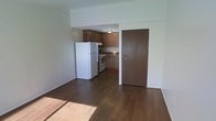 This single-floor space offers a practical and functional layout designed for efficient everyday living. The open-concept design seamlessly connects the living area to the kitchen, creating a sense of continuity throughout the unit. Dark hardwood flooring runs throughout the main living space, adding warmth and a classic aesthetic to the interior.

The kitchen is positioned at the rear of the open area and is equipped with essential appliances, including a full-size white refrigerator and a white electric range. Medium-toned brown cabinetry lines the upper and lower walls of the kitchen, providing ample storage space, while a countertop with a dark surface complements the overall color palette. A ceiling-mounted light fixture provides direct illumination over the kitchen workspace.

Natural light filters into the living area, brightening the light-toned walls and contributing to an airy, welcoming atmosphere. A solid entry door is situated adjacent to the kitchen, offering a clear sense of the unit's layout and flow from the entrance. The overall design is straightforward and practical, making the space well-suited for those seeking a functional and easy-to-maintain living environment.