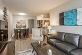 This thoughtfully designed single-floor residence offers a comfortable and functional layout across several well-defined living spaces. The open-concept living and dining area forms the heart of the home, featuring light wood-style flooring, neutral walls, and natural light streaming in through large windows. The living room accommodates a full seating arrangement and is complemented by an adjacent dining area with room for a four-person table and contemporary wall art.

The kitchen is efficiently laid out with dark cabinetry, granite-style countertops, white appliances, and a built-in pantry with open shelving for additional storage. A peninsula with a breakfast bar and an under-counter dishwasher connects the kitchen to the main living space, offering both practicality and casual dining options.

The primary bedroom features plush carpeting, a large window with natural light, and space for a full bedroom suite. A secondary room, carpeted and windowed, offers flexibility as a bedroom or home office. The bathroom is a standout feature, boasting a generous double-sink vanity with dark cabinetry, granite-style countertops, a wide mirror, and wood-style flooring. Throughout, the space maintains a cohesive, contemporary aesthetic with a calm, neutral color palette.
