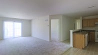 This single-floor space presents a straightforward and functional layout designed to maximize usability. The main living area is generously sized, finished with plush wall-to-wall carpeting in a neutral tone that complements the crisp white walls and ceiling throughout. The open floor plan creates a seamless flow between living spaces, making the area feel spacious and adaptable to various furniture arrangements.

A doorway visible from the main living area leads to an additional room, suggesting a flexible layout suited for multiple uses. Natural light filters in through a window along one wall, brightening the interior and enhancing the airy atmosphere created by the light color palette.

To one side, a partial view of the kitchen area is framed by a decorative white column and half-wall, providing a subtle visual separation while maintaining the open feel of the space. The kitchen features tile flooring, contrasting with the carpeted living area, and includes warm-toned cabinetry. A central air vent is integrated into the wall, indicating a built-in HVAC system. Overall, the space offers a clean, neutral canvas well-suited for personalization.