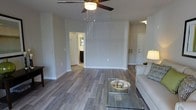This single-floor residence offers a clean, contemporary design defined by a neutral color palette and an open, airy layout. The living area serves as the central hub of the space, finished with wide-plank wood-style flooring in a warm gray tone that runs throughout, adding both visual continuity and a modern aesthetic.

The living room is generously proportioned, with smooth walls painted in a soft gray-white that maximizes the sense of brightness. A ceiling fan with a central light fixture provides both comfort and overhead illumination. The open wall space offers ample flexibility for furniture arrangement, while a glass-top coffee table and a light upholstered sofa add a refined, minimalist character.

Visible from the living area, a hallway leads to what appears to be a bathroom, offering a glimpse of a well-lit vanity and dark cabinetry. On the opposite side, a white interior door provides access to additional areas of the home. A wall-mounted air vent ensures efficient climate control throughout the space.

Natural and artificial light work together to keep the interior feeling fresh and welcoming. The overall ambiance is calm and functional, with a design sensibility that is both practical and modern, making it a versatile space well-suited for everyday living.