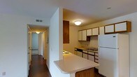This single-floor residence offers a practical and well-organized layout designed for comfortable everyday living. The kitchen serves as a central highlight, featuring a peninsula with a breakfast bar that provides both additional prep space and casual seating. A combination of dark wood and white cabinetry lines the walls, offering generous storage throughout. The kitchen is equipped with essential appliances, including a white refrigerator, a stovetop, and a built-in dishwasher, all centered around a functional sink setup. Overhead flush-mount lighting keeps the space well-illuminated. A half-wall separates the kitchen from the adjacent hallway, which runs the length of the unit and connects to the remaining rooms. The hallway is finished with warm hardwood-style flooring that contrasts with lighter carpet visible in the rooms beyond. White walls throughout give the space a clean, neutral backdrop, while multiple doorways along the corridor hint at a thoughtful room arrangement. HVAC vents are integrated into the ceiling, ensuring climate control throughout. The overall ambiance is practical and move-in ready, with a straightforward floor plan that makes efficient use of the available space.