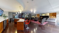 This contemporary single-floor space is designed as a sophisticated communal lounge, combining functional and social areas in an open, well-appointed layout. Rich, dark hardwood flooring flows throughout, anchoring the modern aesthetic and providing a cohesive visual foundation.

At the heart of the space is a curved reception-style bar with warm wood cabinetry and a clean white countertop, accompanied by bold red bar stools. The bar area is outfitted with built-in wine refrigerators and a mounted flat-screen TV, making it a natural gathering hub. Pendant lighting highlights the curved ceiling detail above, adding architectural interest.

The lounge area features a generous seating arrangement with a large sectional sofa, swivel accent chairs, and a floral-patterned area rug in deep rose tones. A striking floor-to-ceiling fireplace with a built-in firewood storage compartment serves as a focal point on one wall, flanked by a wall-mounted television and warm wood built-in shelving with display lighting.

Natural light enters through multiple sets of French doors along the exterior wall, offering views of an outdoor area and creating a bright, welcoming ambiance. Track lighting throughout ensures even illumination across all zones. The overall design balances comfort and style, suited for both relaxation and social interaction.