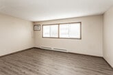 This single-floor unit offers a practical and straightforward layout across several well-defined rooms. The main living area features wood-look laminate flooring, a textured ceiling, and a wall-mounted air conditioning unit, alongside wide windows fitted with horizontal blinds that allow generous natural light to fill the space. Baseboard heating runs throughout, ensuring consistent warmth across the unit.

Adjacent living spaces continue the wood-look flooring and open into one another, with ceiling fans providing additional air circulation. A dedicated area with tile-patterned flooring transitions into the kitchen, which is outfitted with rich, dark-toned wood cabinetry, dark granite-style countertops, and white appliances including a refrigerator, electric range, and microwave. A tile floor in an earthy checkerboard pattern grounds the kitchen space.

The bedrooms feature carpeted flooring in neutral tones, ceiling fans, baseboard heaters, and windows — one offering a view of surrounding trees. The bathroom includes a full tub-shower surround, a toilet, a single-sink vanity with warm wood cabinetry, and matching tile flooring. Throughout the unit, dark-trimmed wood doors and neutral beige walls create a cohesive, understated aesthetic.