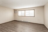 This single-floor unit offers a practical and straightforward layout across several well-defined rooms. The main living area features wood-look laminate flooring, a textured ceiling, and a wall-mounted air conditioning unit, alongside wide windows fitted with horizontal blinds that allow generous natural light to fill the space. Baseboard heating runs throughout, ensuring consistent warmth across the unit.

Adjacent living spaces continue the wood-look flooring and open into one another, with ceiling fans providing additional air circulation. A dedicated area with tile-patterned flooring transitions into the kitchen, which is outfitted with rich, dark-toned wood cabinetry, dark granite-style countertops, and white appliances including a refrigerator, electric range, and microwave. A tile floor in an earthy checkerboard pattern grounds the kitchen space.

The bedrooms feature carpeted flooring in neutral tones, ceiling fans, baseboard heaters, and windows — one offering a view of surrounding trees. The bathroom includes a full tub-shower surround, a toilet, a single-sink vanity with warm wood cabinetry, and matching tile flooring. Throughout the unit, dark-trimmed wood doors and neutral beige walls create a cohesive, understated aesthetic.