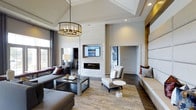 This single-floor space makes an immediate impression with its dramatic vaulted ceilings and refined contemporary design. The main living area serves as the centerpiece, anchored by a sleek, wall-mounted linear fireplace framed in light-toned tile that extends to the ceiling, creating a striking focal point. A wall-mounted display above the fireplace adds a modern, functional element to the design.

The room is illuminated by a combination of recessed lighting and a statement chandelier with an industrial-inspired metal frame, casting warm, inviting light throughout the space. Large windows allow natural light to filter in, enhancing the open and airy atmosphere.

Rich dark hardwood flooring grounds the room, contrasting elegantly with the soft neutral palette of the walls and furnishings. Plush gray sectional seating and coordinated accent chairs are arranged around a sleek coffee table, offering a comfortable yet stylish gathering area. A textured area rug adds warmth and definition to the seating arrangement.

Architectural detailing is evident throughout, from the coffered ceiling elements to the wood-paneled accent wall visible through a connected passage. Built-in bench seating along one wall provides additional functionality while maintaining the clean aesthetic. The overall ambiance is one of polished sophistication, blending modern design with inviting comfort.