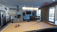 Located at 2000 Front Street in Summerville, South Carolina, this single-floor community clubhouse offers a well-appointed gathering space designed for both recreation and socializing. The open-concept layout seamlessly connects multiple functional zones, creating an inviting and versatile environment.

At the heart of the space is a dedicated billiards area, complete with a full-size pool table and a striking modern pendant light fixture that anchors the room. Adjacent to this is a fully equipped kitchen featuring white cabinetry, stainless steel appliances including a professional-style range hood, and a granite-topped island with bar seating — ideal for entertaining.

A statement accent wall finished in a bold herringbone tile pattern serves as a dramatic focal point, framing a wall-mounted flat-screen television and creating a defined lounge and dining zone. The dining area accommodates a large group around a substantial table with upholstered chairs, making it well-suited for community events and gatherings.

Large windows flood the space with natural light and offer views of the surrounding outdoor area, enhancing the open, airy ambiance. The overall design blends contemporary finishes with functional amenities, delivering a polished and comfortable space for residents and guests alike.