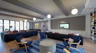 This contemporary single-floor space functions as a well-appointed communal lounge, designed with both comfort and style in mind. The interior showcases a cohesive blue color palette, anchored by plush navy blue sofas and accent chairs with warm wood frames, arranged to encourage both intimate conversation and casual gathering. A bold black-and-white striped cylindrical ottoman serves as a striking centerpiece atop a patterned blue area rug, adding a dynamic visual element to the seating arrangement. Overhead, globe pendant lights hang at varying heights from a coffered ceiling, casting a warm, inviting glow throughout the space. Recessed lighting complements the pendants, ensuring the room is well-lit at all times. A wall-mounted flat-screen TV is positioned above a sleek, built-in white credenza, creating a dedicated media area that integrates seamlessly into the design. Colorful artwork adorns the neutral gray walls, adding personality and visual interest. Large windows along one side of the room allow natural light to filter in, enhancing the open and airy atmosphere. The light wood flooring grounds the space with a natural warmth that balances the modern furnishings. Overall, this lounge offers a sophisticated and functional environment suited for relaxation and social engagement.