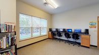 This single-floor facility is thoughtfully designed to serve as a community learning and technology hub. The space is organized into distinct functional areas, each tailored to support education and digital access.

The computer labs are the centerpiece of the facility, featuring multiple workstations equipped with desktop computers arranged along built-in countertops with warm wood cabinetry. The primary lab offers a generous row of workstations bathed in natural light from large windows fitted with horizontal blinds, providing pleasant outdoor views of the surrounding neighborhood. A secondary workstation area near the entrance offers additional desktop setups, also framed by French-style wood doors and a wall-mounted mirror that adds depth to the space.

A dedicated study room provides a quieter environment for individual or small-group learning, complete with a table and chairs, a whiteboard for instruction or brainstorming, and a bookshelf stocked with reading materials. Windows on two walls fill the room with natural light, fostering a calm and focused atmosphere.

Throughout the facility, neutral-toned carpet, cream-colored walls, and consistent warm wood trim create a cohesive, approachable aesthetic. Overhead fluorescent lighting ensures all areas are well-lit. The layout is open and accessible, making it a functional and welcoming space for community members seeking educational resources and technology support.