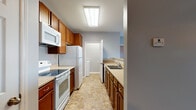 This single-floor unit offers a thoughtfully laid-out living space with a cohesive neutral color palette of soft blue-gray walls, white trim, and warm beige carpeting throughout the main living and bedroom areas. The galley-style kitchen is well-equipped, featuring warm-toned wood cabinetry, light countertops, white appliances including a range, built-in microwave, refrigerator, and dishwasher, along with a double basin sink. Stone-look tile flooring runs through the kitchen and hallway areas, adding durability and visual interest. An overhead flush light fixture brightens the kitchen corridor, which connects to the rest of the unit with ease. The open living area is spacious and inviting, anchored by a ceiling fan and access to an outdoor balcony through a glass door flanked by large windows that invite natural light. A half-wall separates the living space from the kitchen, creating a sense of openness while maintaining functional distinction. The bedrooms are comfortably sized, each featuring plush carpeting, ceiling fans, and windows with blinds that allow for natural light and privacy. The two full bathrooms are finished with stone-look tile flooring, warm wood vanities, and full tub-shower combinations with chrome fixtures, offering clean and functional design.