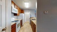 This single-floor unit offers a thoughtfully laid-out living space with a cohesive neutral color palette of soft blue-gray walls, white trim, and warm beige carpeting throughout the main living and bedroom areas. The galley-style kitchen is well-equipped, featuring warm-toned wood cabinetry, light countertops, white appliances including a range, built-in microwave, refrigerator, and dishwasher, along with a double basin sink. Stone-look tile flooring runs through the kitchen and hallway areas, adding durability and visual interest. An overhead flush light fixture brightens the kitchen corridor, which connects to the rest of the unit with ease. The open living area is spacious and inviting, anchored by a ceiling fan and access to an outdoor balcony through a glass door flanked by large windows that invite natural light. A half-wall separates the living space from the kitchen, creating a sense of openness while maintaining functional distinction. The bedrooms are comfortably sized, each featuring plush carpeting, ceiling fans, and windows with blinds that allow for natural light and privacy. The two full bathrooms are finished with stone-look tile flooring, warm wood vanities, and full tub-shower combinations with chrome fixtures, offering clean and functional design.
