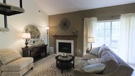 This well-appointed two-floor home offers a thoughtfully designed interior that balances comfort and style. The living room serves as the heart of the space, anchored by a striking wood-mantled fireplace with a classic white surround and a rich, earthy accent wall that adds depth and warmth to the room. A decorative medallion wall piece above the mantel serves as a focal point, complementing the refined aesthetic. Natural light filters in through a large window dressed with elegant cream curtains, brightening the space and creating an airy feel. The living area is generously sized, accommodating a plush sofa with accent pillows, a round patterned area rug, and a sleek circular coffee table — all contributing to a cohesive and inviting atmosphere. A console table with a decorative round mirror and accent lamp adds a stylish vignette along one wall. The neutral tones of taupe, cream, and warm brown carry throughout, evoking a sense of calm and sophistication. Spanning two floors, this home provides ample space for both everyday living and entertaining, with a layout that encourages a smooth and natural flow between levels.