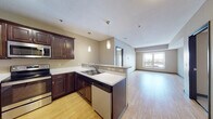 This single-floor residential unit offers a thoughtfully designed open-concept layout that seamlessly connects the kitchen and living area. The kitchen is a standout feature, showcasing rich dark-stained cabinetry, stainless steel appliances including a refrigerator, electric range, and built-in microwave, complemented by light countertops and a double-basin sink. A peninsula with a granite-topped breakfast bar and pendant lighting above adds both functionality and style. Light hardwood-style flooring runs throughout the main living and kitchen areas, enhancing the bright, open feel.

The living area extends naturally from the kitchen, bathed in natural light from a large window offering exterior views. Two carpeted bedrooms provide comfortable retreats, each featuring windows with outdoor views and generous closet space with light wood bifold doors.

The unit includes two bathrooms. The primary bathroom features a floating vanity with dark cabinetry, a white countertop, a large mirror, and a tall linen tower for ample storage. The second bathroom offers a single-sink vanity with matching dark cabinetry and a full tub-shower combination with tile flooring.

A dedicated utility corridor houses a stacked washer and dryer unit alongside additional storage and closet access, adding everyday convenience to this well-rounded living space.