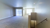 This single-floor residence offers a clean, open-concept layout designed for comfortable everyday living. The main living area is generously proportioned, featuring wall-to-wall carpeting that adds warmth and softness underfoot. Large sliding glass doors dominate one wall, flooding the space with natural light and providing direct access to an outdoor balcony, making it an inviting spot for relaxing or entertaining. A pass-through window connects the living area to an adjacent room, enhancing the open flow between spaces and adding a practical architectural touch. Recessed lighting in the ceiling provides additional illumination, complementing the abundance of daylight. Baseboard heating units are positioned along the exterior walls, ensuring consistent comfort throughout the cooler months. A stairwell with a handrail is accessible from the main living area, leading to a lower entry level with a door visible at the base. The neutral color palette throughout — from the soft beige walls to the light carpeting — creates a calm, versatile backdrop that accommodates a wide range of furnishing styles. Overall, the space presents a straightforward, functional design with ample room to personalize.
