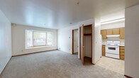 This single-floor unit offers three bedrooms and one bathroom within a well-organized layout. The living area is carpeted and anchored by a wide bay window that frames views of surrounding greenery, allowing natural light to fill the space throughout the day. White walls and smooth ceilings keep the interior feeling bright and clean.

The kitchen is positioned just off the main living area and features light wood cabinetry, white appliances including a refrigerator and range, and tile flooring — providing a functional workspace that transitions smoothly into the living space.

The spacious primary bedroom is notably generous relative to the overall footprint of the unit, offering ample room for furnishings. It features direct access to the full bathroom, which includes a tub/shower combo, a vanity with wood-trimmed cabinetry, tile flooring, and clean white fixtures. The two additional bedrooms are carpeted and each include windows that bring in natural light, with one room featuring a built-in closet with a hanging rod for convenient storage.

Throughout the unit, warm wood-trimmed door frames and baseboards provide consistent detailing. Additional storage is available via a built-in closet with shelving visible from the main living area, adding to the unit's practical functionality.