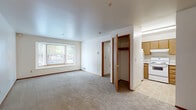 This single-floor unit offers three bedrooms and one bathroom within a well-organized layout. The living area is carpeted and anchored by a wide bay window that frames views of surrounding greenery, allowing natural light to fill the space throughout the day. White walls and smooth ceilings keep the interior feeling bright and clean.

The kitchen is positioned just off the main living area and features light wood cabinetry, white appliances including a refrigerator and range, and tile flooring — providing a functional workspace that transitions smoothly into the living space.

The spacious primary bedroom is notably generous relative to the overall footprint of the unit, offering ample room for furnishings. It features direct access to the full bathroom, which includes a tub/shower combo, a vanity with wood-trimmed cabinetry, tile flooring, and clean white fixtures. The two additional bedrooms are carpeted and each include windows that bring in natural light, with one room featuring a built-in closet with a hanging rod for convenient storage.

Throughout the unit, warm wood-trimmed door frames and baseboards provide consistent detailing. Additional storage is available via a built-in closet with shelving visible from the main living area, adding to the unit's practical functionality.